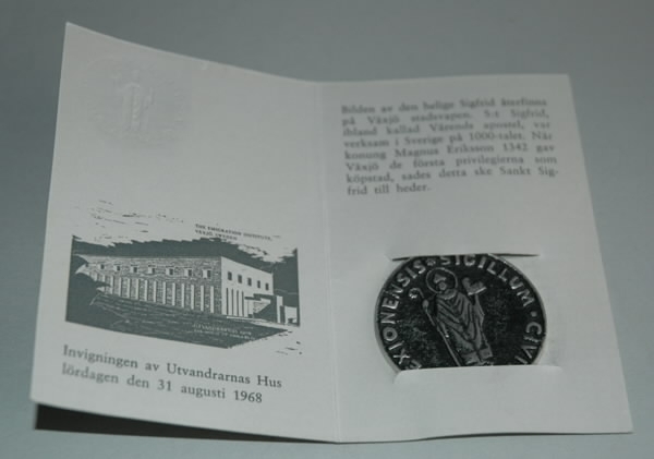 Minnespeng av vitmetall. På framsidan sankte Sigfrids bild och texten SIGILLUM CIVITATIS WEXIONENSIS mot svartanlöpt grund.