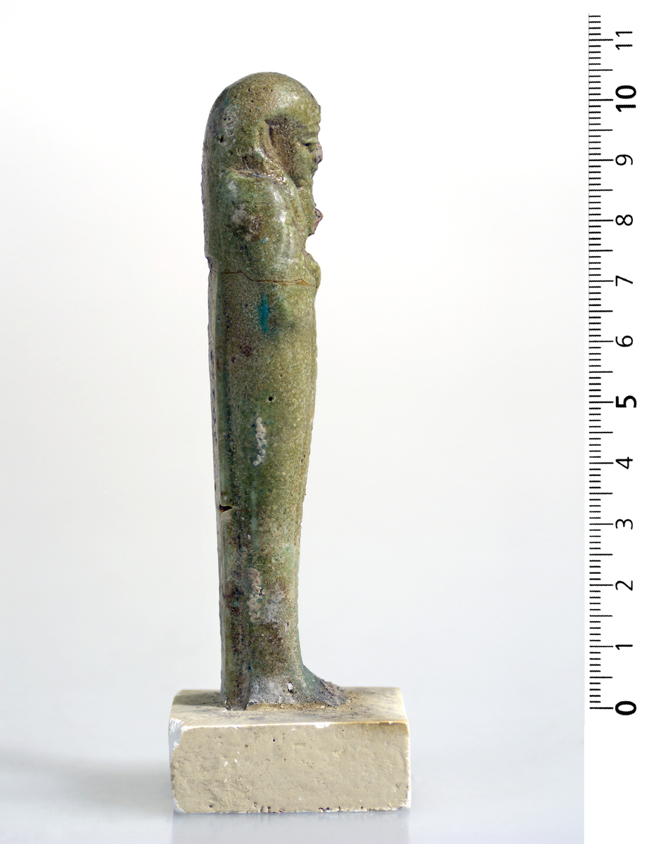 Ushabti i mørk grønn fajanse med hieroglyfer foran. 10,5 cm høy.
Fajanse