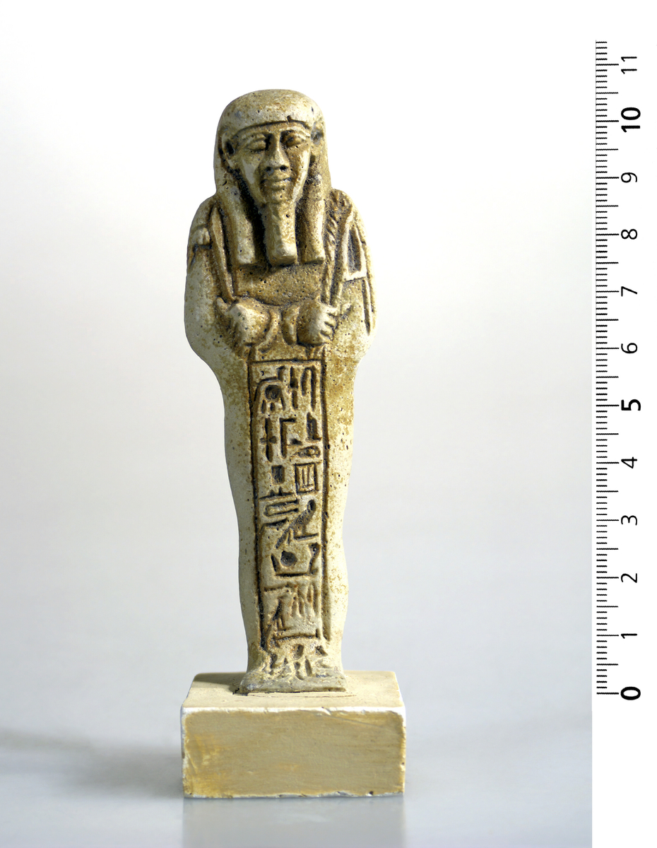 Ushabti i hvit-gul fajanse med tydelige hieroglyfer foran. Pene trekk. 10,5 cm høy.
Fajanse