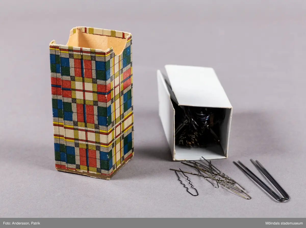 En liten rektangulär ask som innehåller hårnålar. Asken har ett lock som öppnas uppåt. Asken är dekorerad med ett geometriskt mönster som är indelat i rutor och ränder. Askens mönster liknar en Mondrian målning.