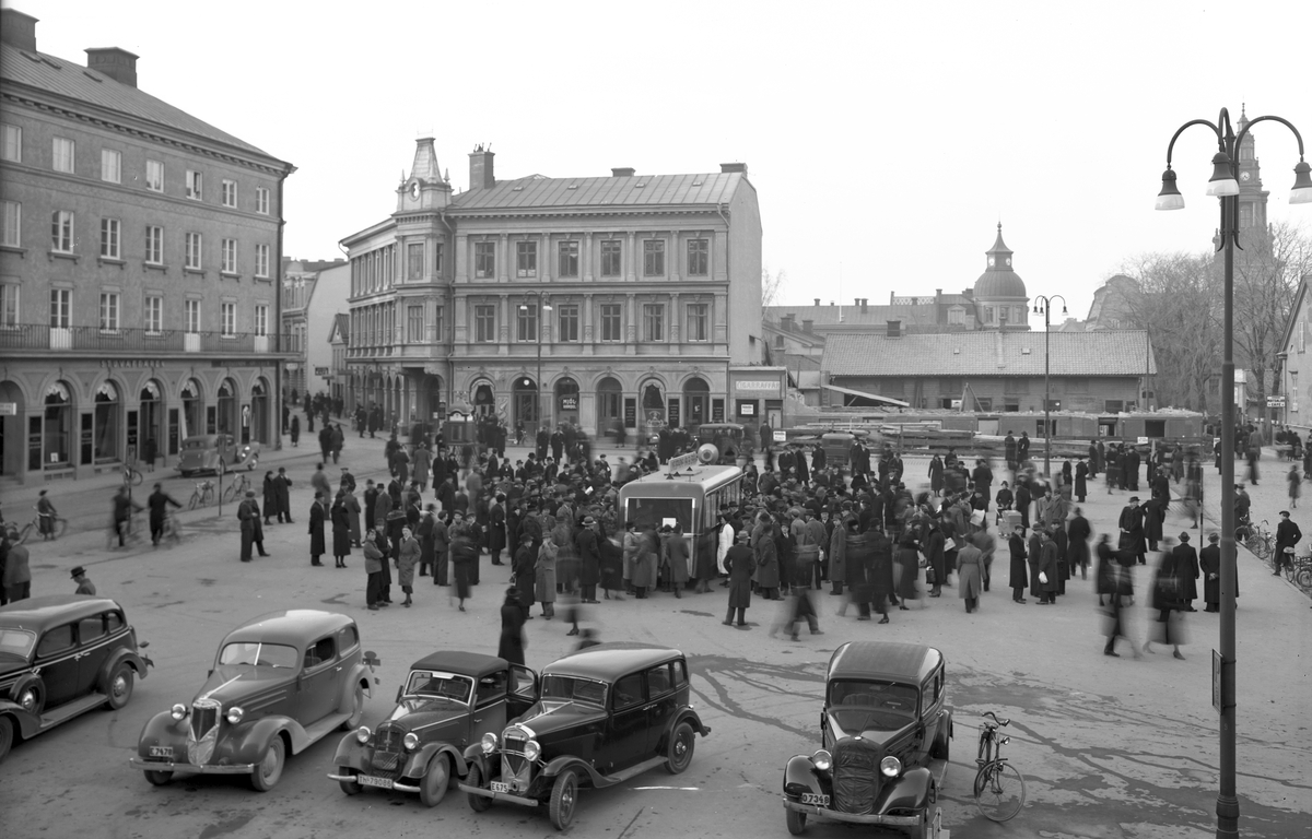 Illustrativ vy över Trädgårdstorget i Linköping där människorna i stunden samlats vid Linguaphones demonstration av sina språkkurser via grammofonskivor. Bilden kan genom den pågående rivningen av Mörnerska / Wentzellska gården vid torgets norra del dateras till 1930-talets sista år. Under decenniets sista år kom även fasaden till Nylanderska huset till vänster att förenklas för att anpassas till det nya, blivande Trädgårdsföreningens hus.