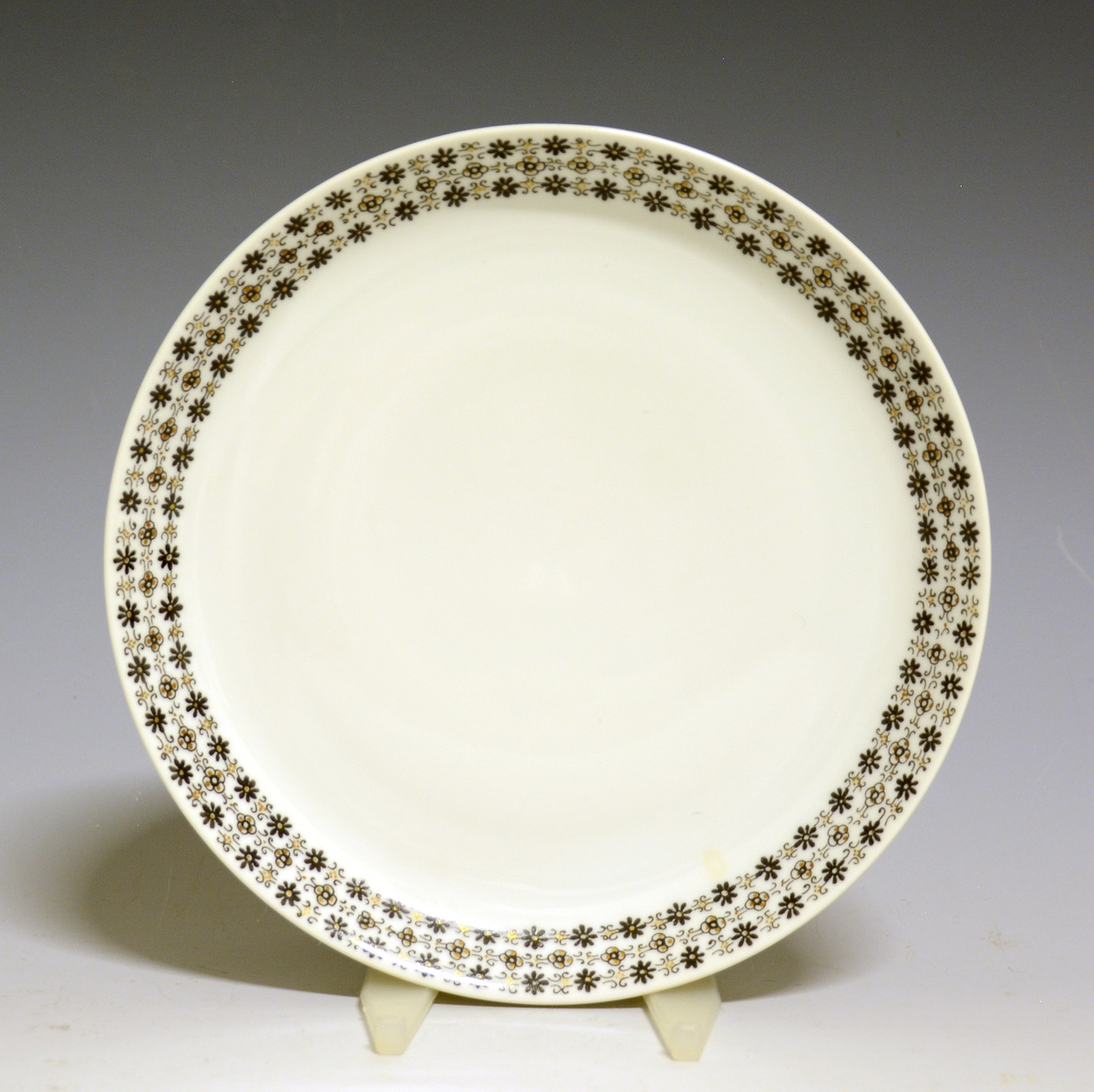 Asjett av porselen med hvit glasur og bord med småblomstret trykkdekor i sort og gull langs kanten.
Modell: Petita (eller variant av)
