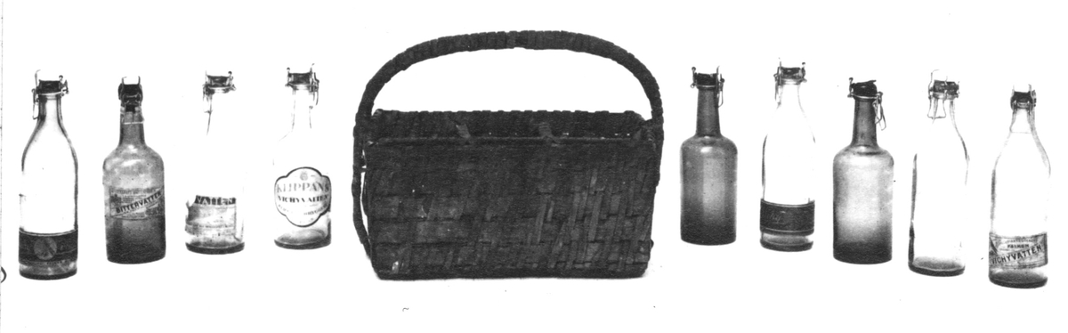 a) Flätad korg av säv. Längd: 33 cm, bredd: 25 cm, höjd: 16 cm.Rektangulär med handtag och sex fack, 10 x 11 cm. b-c) Flaska, glas, grön. Etikett saknas. Höjd: 23 cm, diameter: 7 cm.Patentkork av porslin. d) Flaska, glas, grön.Höjd: 23 cm, diameter: 7 cm. Patentkork av porslin. Rektangulär etikett, rosa botten, svart text: "APOTEKARNES VATTENFABRIK - BITTERVATTEN". Även etikett runt halsen. e) Flaska, glas, grön. Höjd: 23 cm, diameter: 7 cm. Patentkork av porslin. Etikett: Vit botten, röd och svart text: "KLIPPANS VICHYVATTEN." (Se etikettens form på scannad bild.) f) Flaska, glas, ljusgrön. Höjd: 23 cm, diameter: 7 cm.Patentkork av porslin. Trasig rektangulär etikett, gul botten, svart text:  "- VATTEN, AKTIEBOLAGET NYA BRYGGERIET I BORÅS.""g) Flaska.Höjd: 24 cm, diameter: 7 cm. Etikett saknas. Av glas. Ljusgrön. Patentkork av porslin. h) Flaska. Höjd: 24 cm, diameter: 7 cm. Av glas. Ljusgrön. Patentkork av porslin. Rektangulär etikett, gul botten med svart text: "Bryggeriaktiebolaget FALKEN, Vichyvatten." i-j) Flaska. Höjd: 24 cm, diameter: 7 cm. Av glas. Ljusgrön. Patentkork av porslin. Rektangulär etikett, svart botten med guld. Röd text: "Vichyvatten, Bryggeriaktiebolaget FALKEN, Falkenberg."