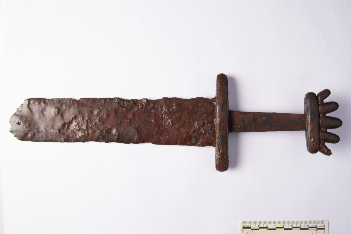 "Stykker av tveegget sverd av typen Jan Petersen l. c., fig. 106 (type O). Klingen er bevart til en lengde av 78 cm., men odden mangler. Klingens bredde innerst ved hjaltet er 5,6 cm. Der er spor efter tvunnet sølvtråd mellem knappens tunger, derimot kan ikke ses spor efter sølvbelegg på hjaltene. Underhjalt 10,9 cm. l., overhjalt 6,6 cm. l." Ny beskrivelse etter revisjon: Tretten fragmenter av tveegget sverd av jern med sølvtråder på hjaltene, Petersens type O. Et stort fragment av håndtak og øvre del av klingen, åtte større fragmenter av nedre del av klingen, fem av disse fastrustet med deler av spydspissen b, pilspisser d, skjoldbulen e og hesteutstyr, samt fire mindre klingefragmenter. Sverdhåndtaket er velbevart med knapp med fem tunger, den ene ytterste av disse noe skadet. Vertikalt mellom tungene har det gått tredobbel sølvtråd, en glatt midttråd og tvunnede sidetråder. Mellom en av tungene mangler tråden nå. Knappens sth: 25, stl: 72 mm, stt: 12 mm. Overhjaltet har avrundede kanter og fure nederst med delvis bevart, horisontal, tvunnet sølvtråd. Overhjalt sth: 12 mm, stl: 67 mm, stt: 17 mm. Grepet har br. øverst 18 mm, nederst 25 mm og stl: 84 mm. Underhjaltet har avrundete kanter og fuer øverst og nederst med delvis bevart, horisontal, tvunnet sølvtråd. I ene enden er underhjaltet noe oppsprukket. Underhjalt sth: 14 mm, stl: 110 mm, stt: 20 mm. Klingefragmentet under grepet er noe skadet langs eggene og har stl: 235 mm, br. øverst 55 mm, br. nederst: 50 mm. De øvrige åtte store klingefragmentene har br. øverst 51 mm, br. nederst 33 mm, og danner med det øvre klingefragmentet en sammenhengende stl. på ca. 780 mm. Odden mangler. De store fragmentene har stl. hhv. 55 mm, 26 mm, 125 mm, 72 mm, 68 mm, 109 mm, 75 mm og 84 mm. Sverdets omtrentlige samlede stl. 920 mm.