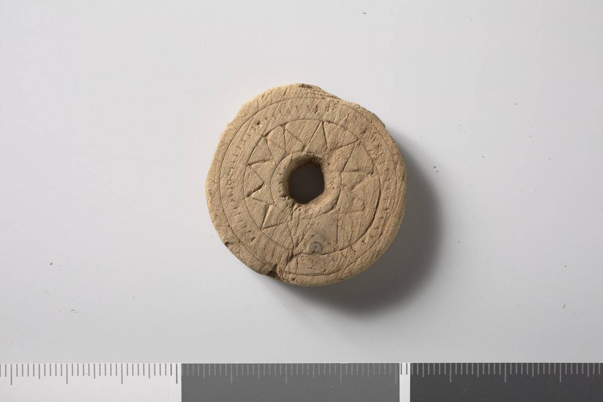 Spinnehjul av tre. Flat, sirkulær skive med hull (0,9 cm). Utskåret sikksakkdekor som er posisjonert "riktig" med tanke på hullet. Diam: 3,8 cm, Stt: 0,9 cm. 