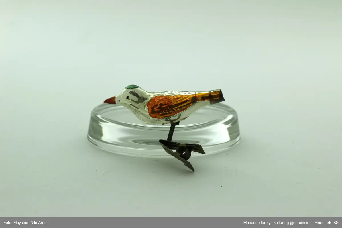 To små juletrefugler laget av plast og med metallfeste. Fuglene har en blank aluminiumsfarge, med påmalte farger. Vinngene er gullfargede, nebbet er rødt og på toppen av hodet er det påmalt en grønn sirkel.