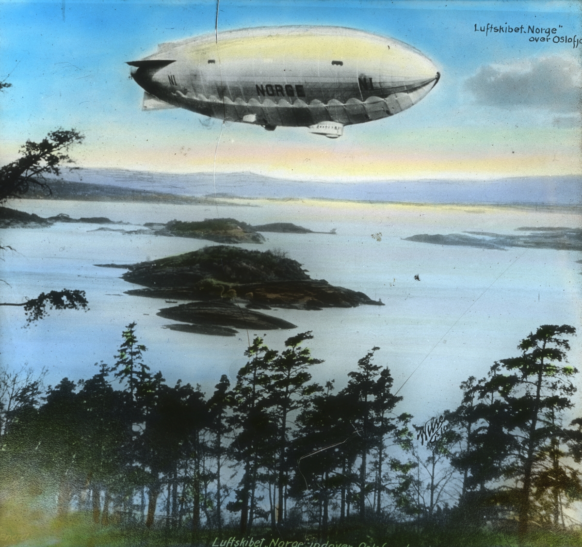 Håndkolorert dias. Luftskipet "Norge" over Oslofjorden fotografert fra Ekeberg i 1926. "Norge" var et mellomstort, halvstivt luftskip bygd av den italienske ingeniøren Umberto Nobile. Roald Amundsen kjøpte det i 1926 og gav det navnet Norge. Luftskipet ble brukt under et fremstøt mot jordens geografiske nordpol under ledelse av Amundsen og i regi av Norsk Luftseiladsforening. Den 29. mars 1926 forlot skipet Roma med Nobile som fører. Landing i Oslo fant sted 14. april ved en spesialbygd fortøyningsmast på Ekebergsletta. Nordpolen ble nådd natten 12. mai. 