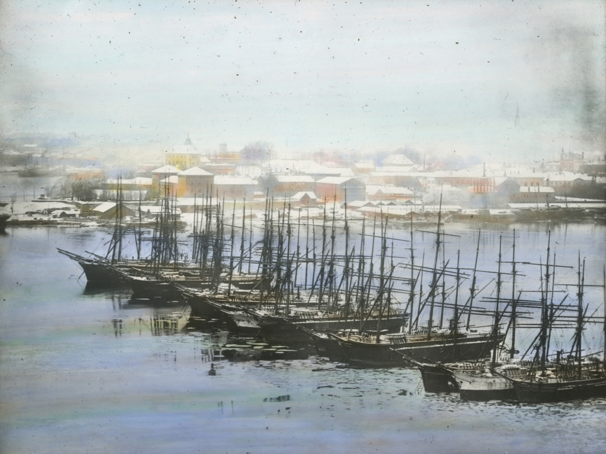 Håndkolorert dias. Seilskuter ligger fortøyd i havnebassenget i Oslo havn vinteren 1901. I bakgrunnen sees Akershus festning og sentrum.