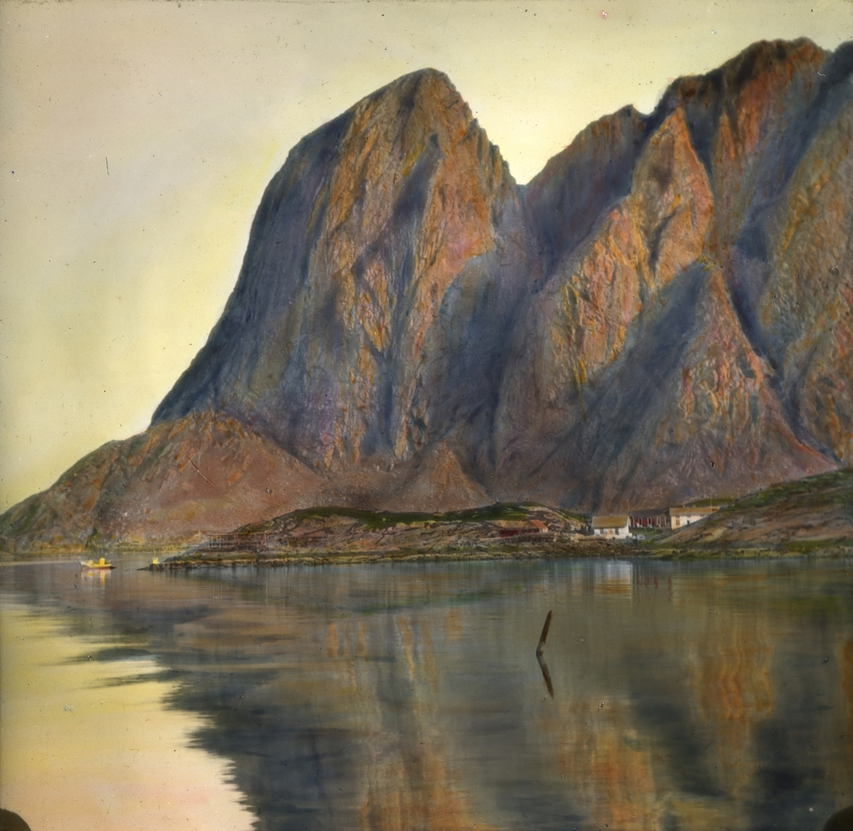 Håndkolorert dias. Midnatt i Reine. Bratte fjellsider ses i bakgrunnen. Havet ses i forgrunnen. Tre-fire gårdsbygninger og en båt ligger ned til vannet.
