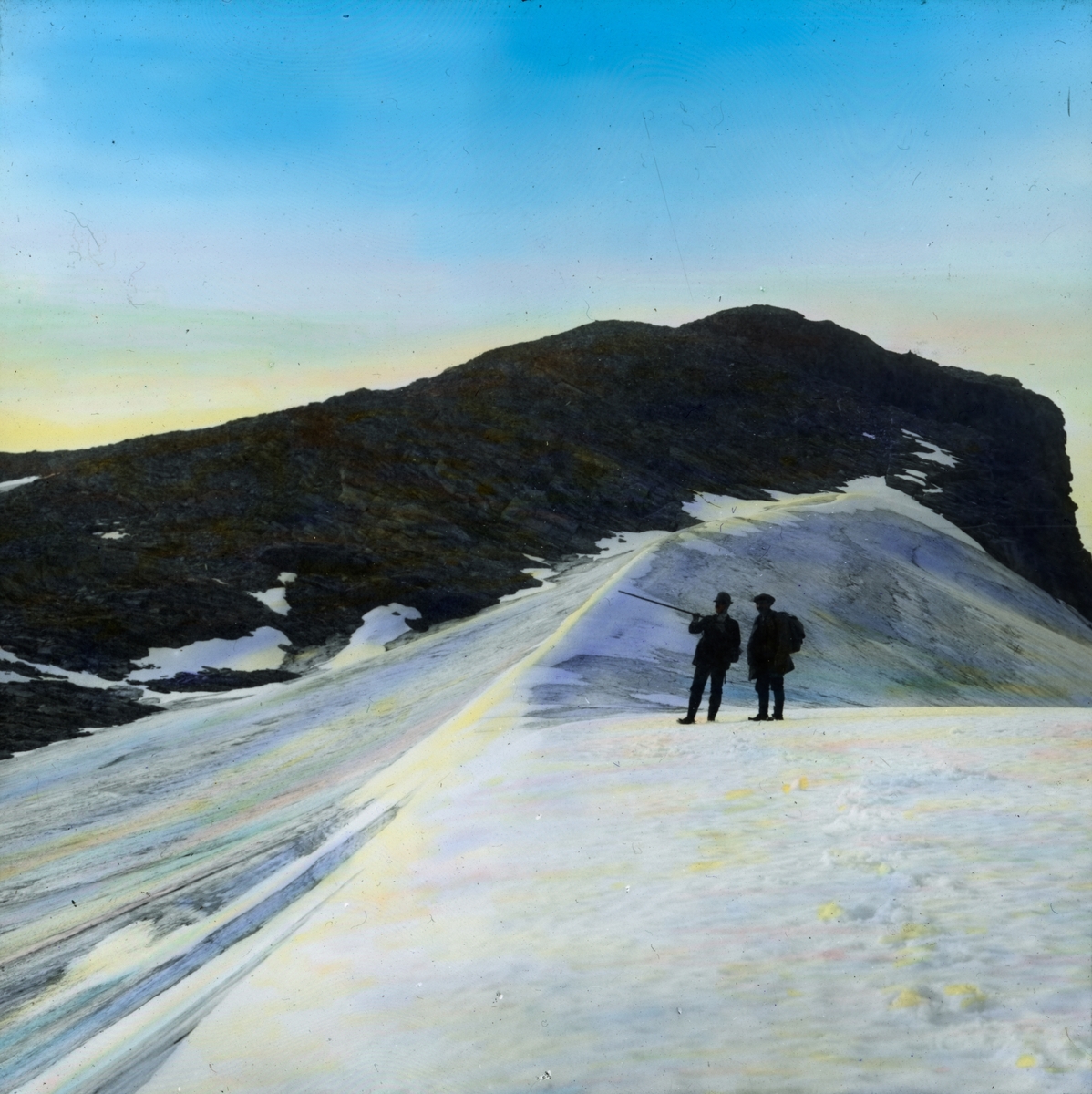 Håndkolorert dias. To menn på toppen av isbreen Vuottarest. En har en ryggsekk, mens den andre peker med en stokk.