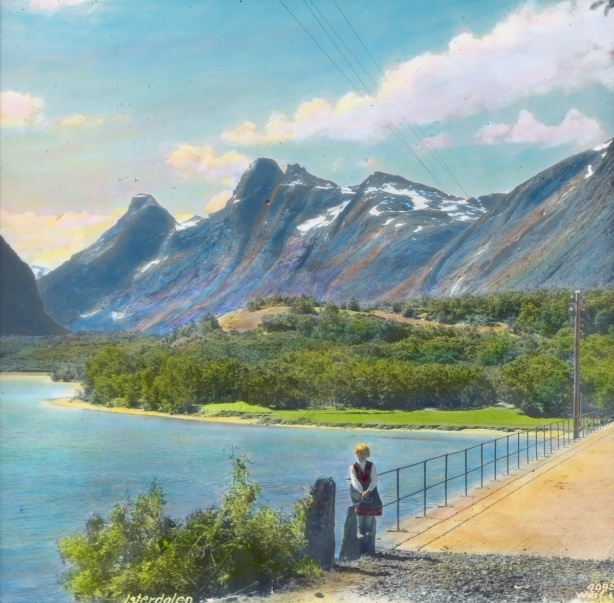 Håndkolorert dias. En jentekledd i folkedrakt sitter på en steinstøtte ved en bro i Isterdalen. I bakgrunnen sees elva Ister og bratte fjell.