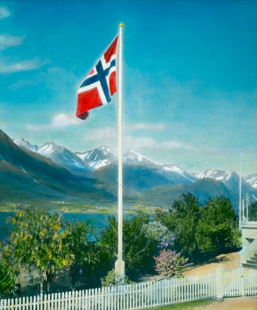 Håndkolorert dias. Et heist norsk flagg på innsiden av et hvitt stakittgjerde. Bak flagget vokser hagebusker og trær. I bakgrunnen ses høye fjell og en fjord.