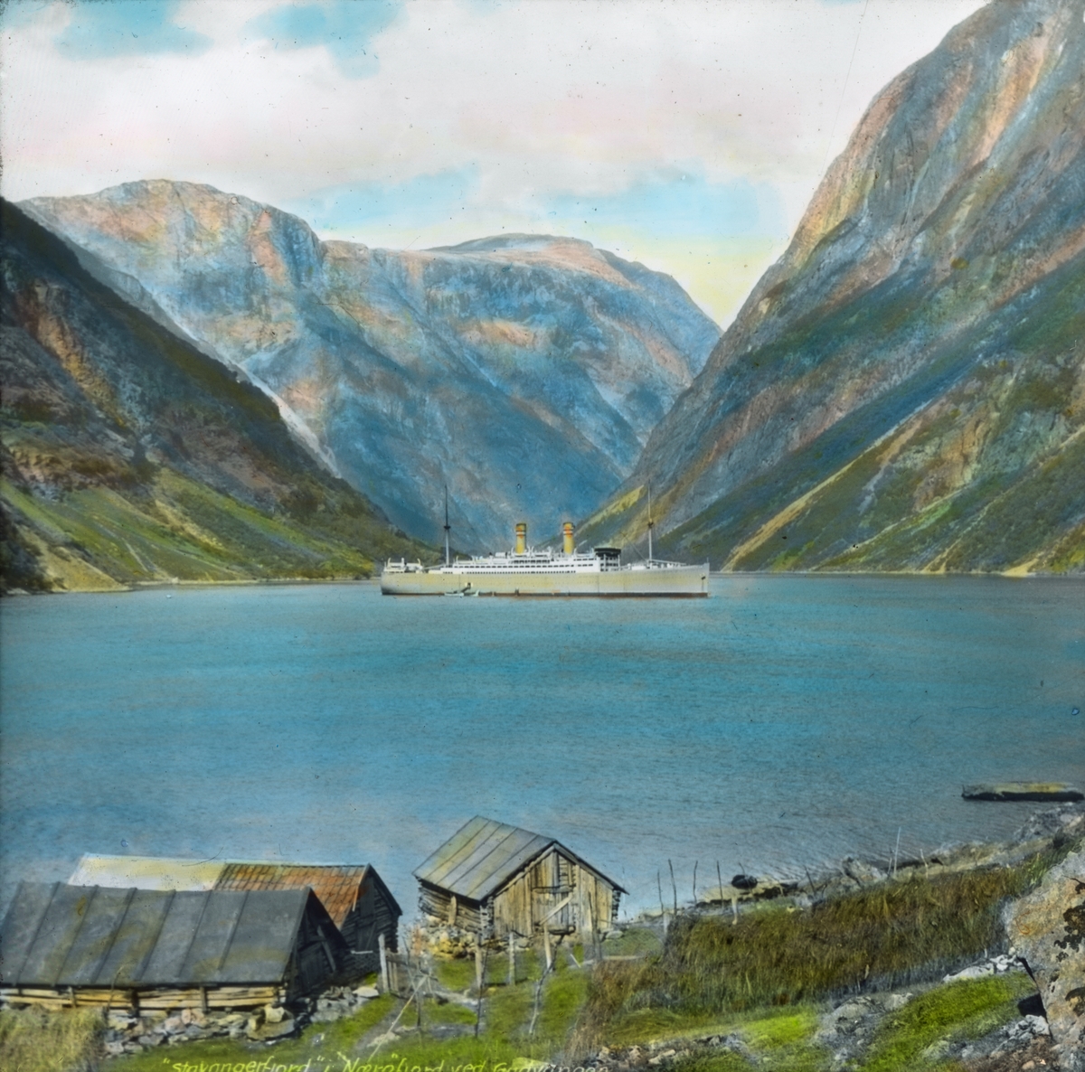 Håndkolorert dias. Et passasjerskip seiler på Nærøyfjorden. I forgrunnen ses en fjellgård. I bakgrunnen ses fjell.