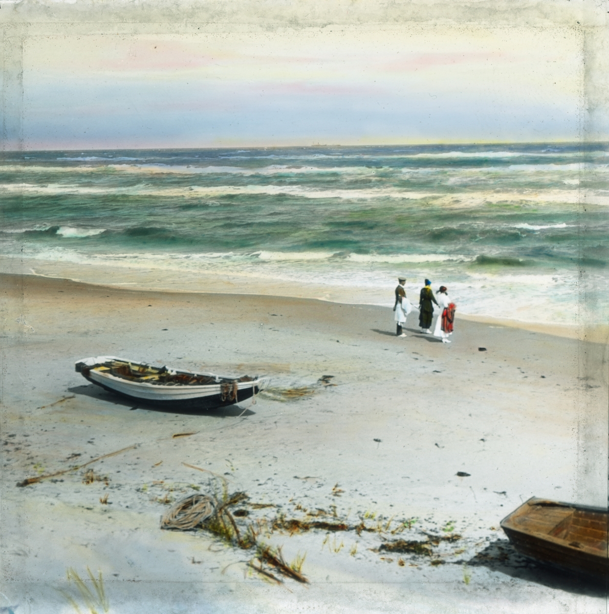 Håndkolorert dias. To kvinner og en mann ser utover havet på en sandstrand. I forgrunnen ligger to robåter i sanden.