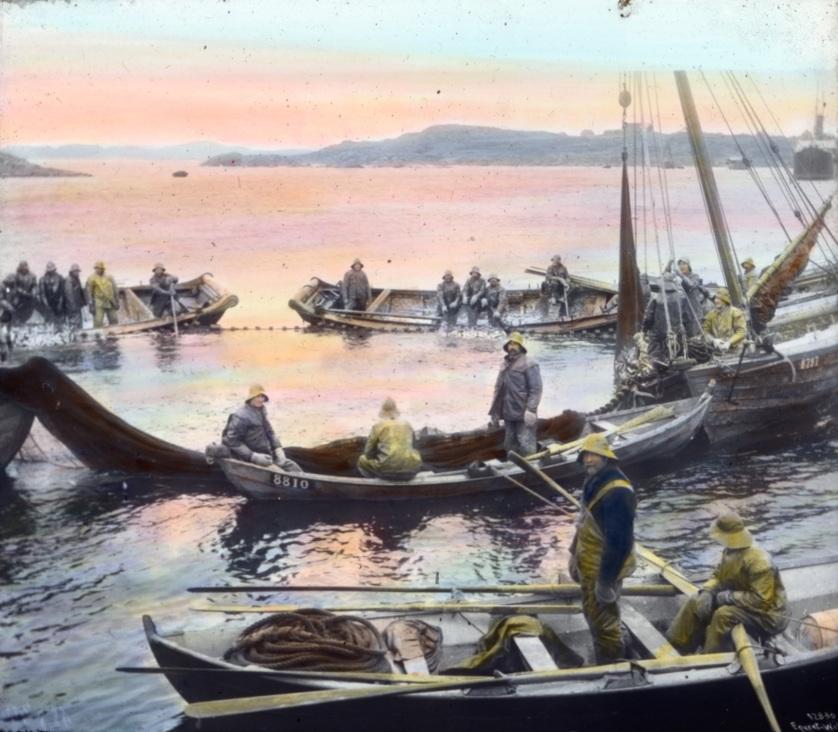 Håndkolorert dias. Flere robåter og en fiskeskøyte ligger omkranset en snurpenot.