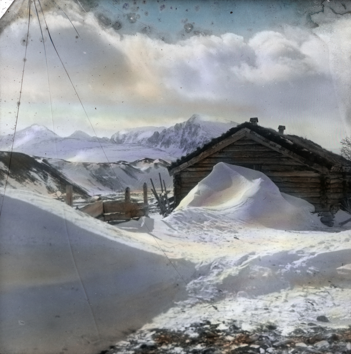 Håndkolorert dias. En laftet tømmerstue sees bak en høy snøskavl. I bakgrunnen ligger snødekte fjell.