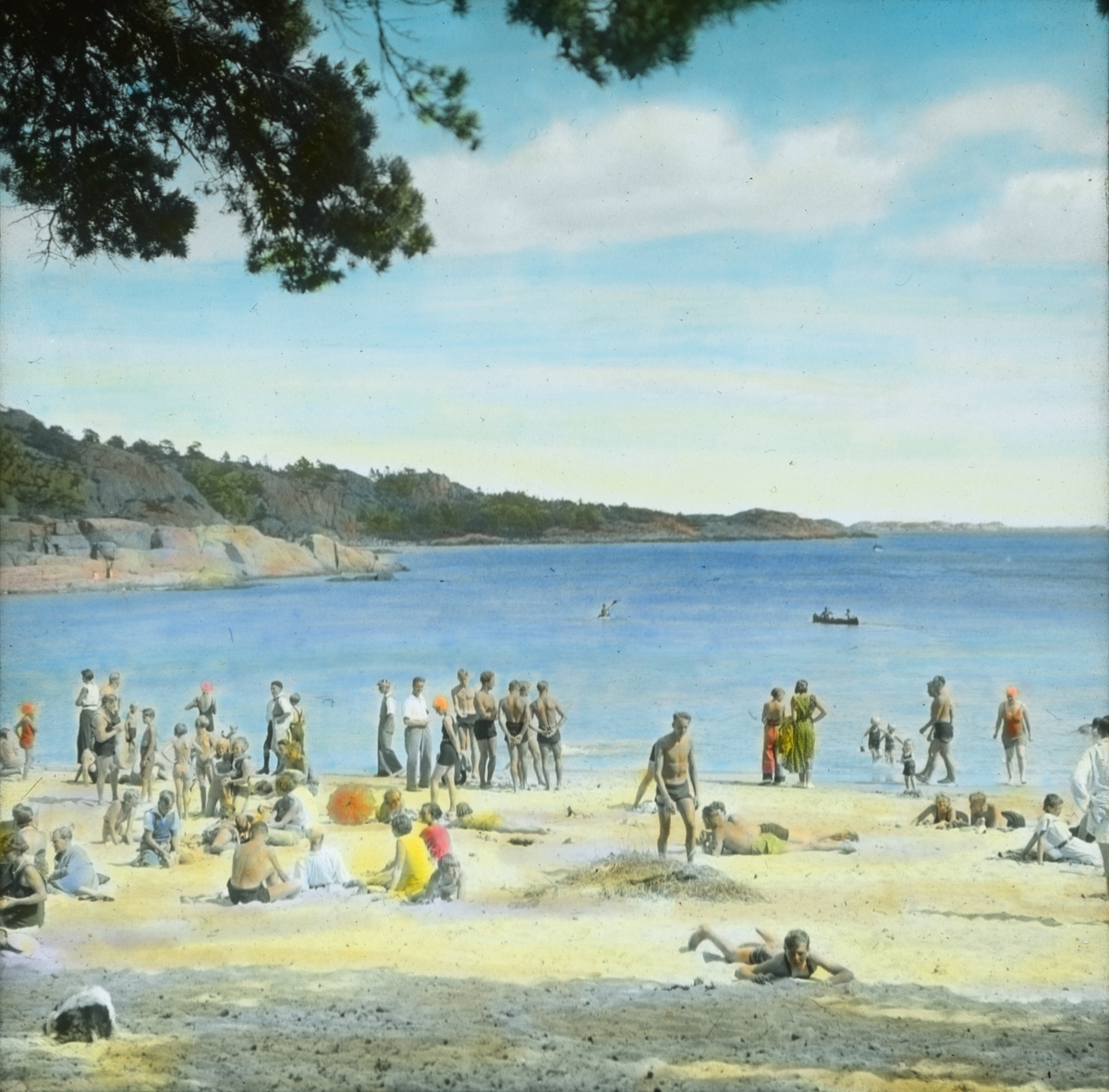 Håndkolorert dias. Kvinner, menn og barn bader og soler seg på stranden.