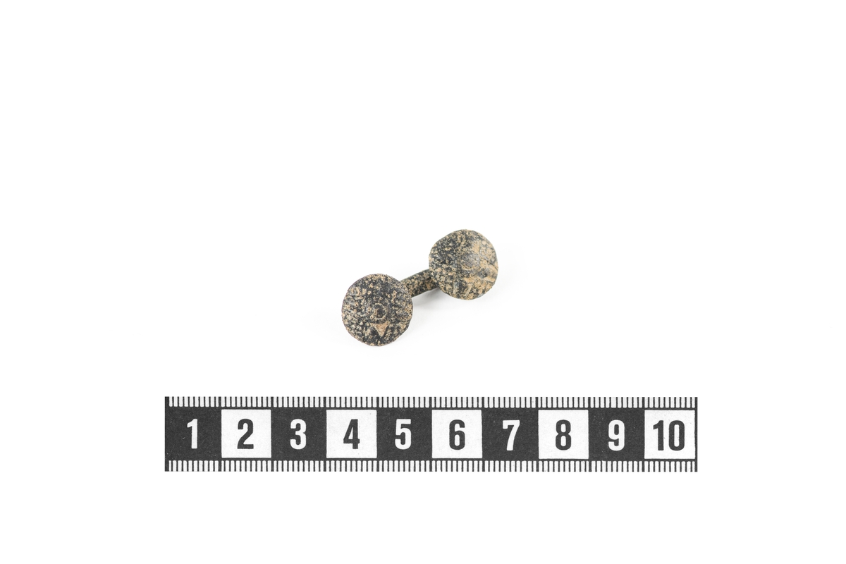 Manschettknapp möjligen av silver. Knapp bestående av en stång med två runda knappar i båda ändar. Knappar är dekorerade med geometriska mönster.