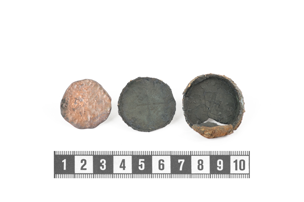 Kopparmynt och fragment av rund cement som omger myntet, en botten (två delar) och ett lock. Ytan på själva myntet är ojämnt, men ett kors är fortfarande svagt synligt på ena sidan. Myntets avtryck är bättre bevarat på insidan av cementet. På huvudsidan verkar det finnas en korsning mellan ett A och ett F. På andra sidan ses två korslagda pilar med bokstäverna R respektive S i motsatta ytor.