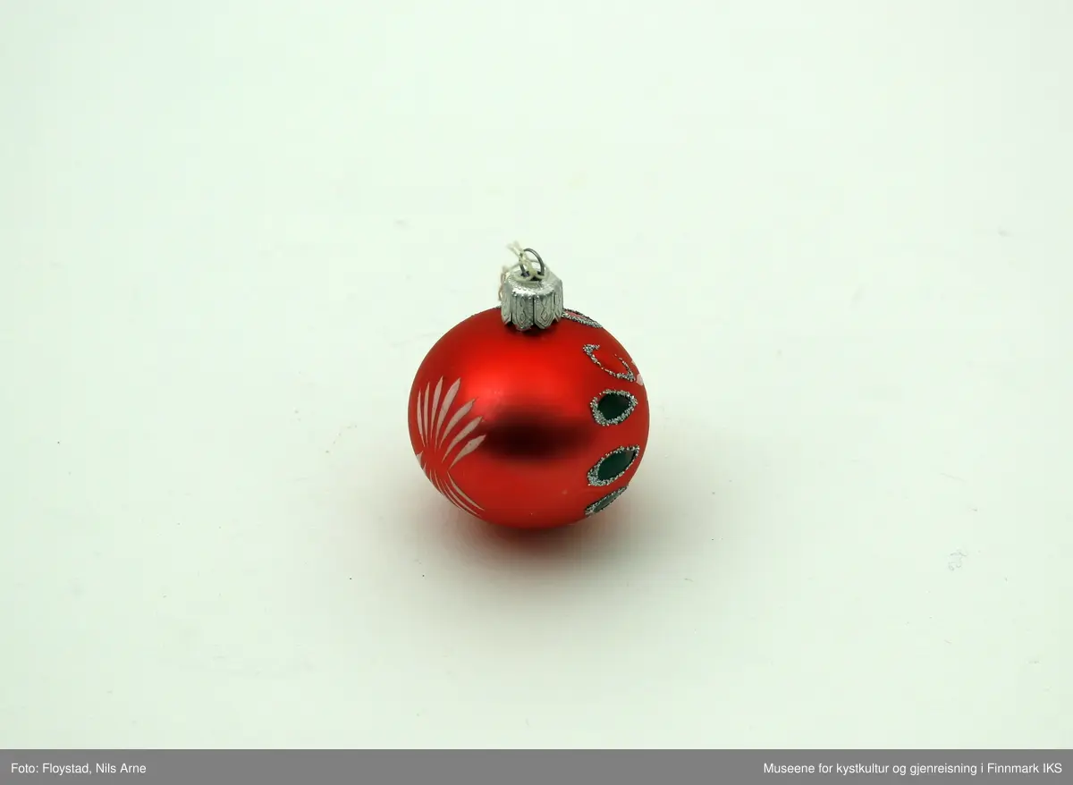 En liten julekule i hard plast med en halvmatt rød/mørkrosa farge med dekor. Dekoren består av påmalte hvite striper på begge sider av kulen og har et blomsterlignende mønster. I tillegg er det dråpeformet dekor malt rød og grønn med glitterkant som går vertikalt rundt kulen, og danner tilsammen en linje. Julekulen har et oppheng av metall med en hvit bomullstråd. Opphenget har gravert mønsterdekor.