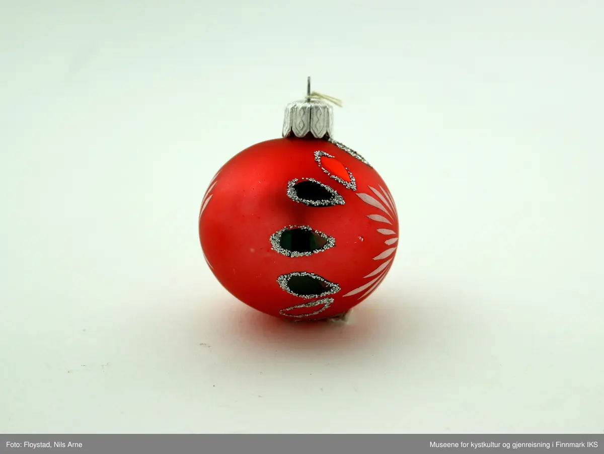 En liten julekule i hard plast med en halvmatt rød/mørkrosa farge med dekor. Dekoren består av påmalte hvite striper på begge sider av kulen og har et blomsterlignende mønster. I tillegg er det dråpeformet dekor malt rød og grønn med glitterkant som går vertikalt rundt kulen, og danner tilsammen en linje. Julekulen har et oppheng av metall med en hvit bomullstråd. Opphenget har gravert mønsterdekor.