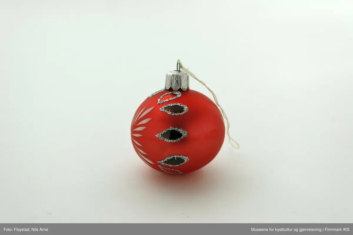 En liten julekule i hard plast med en halvmatt rød/mørkrosa farge med dekor. Dekoren består av påmalte hvite striper på begge sider av kulen og har et blomsterlignende mønster. I tillegg er det dråpeformet dekor malt rød og grønn med glitterkant som går vertikalt rundt kulen, og danner tilsammen en linje. Julekulen har et oppheng av metall med en hvit bomullstråd. Opphenget har gravert mønsterdekor.