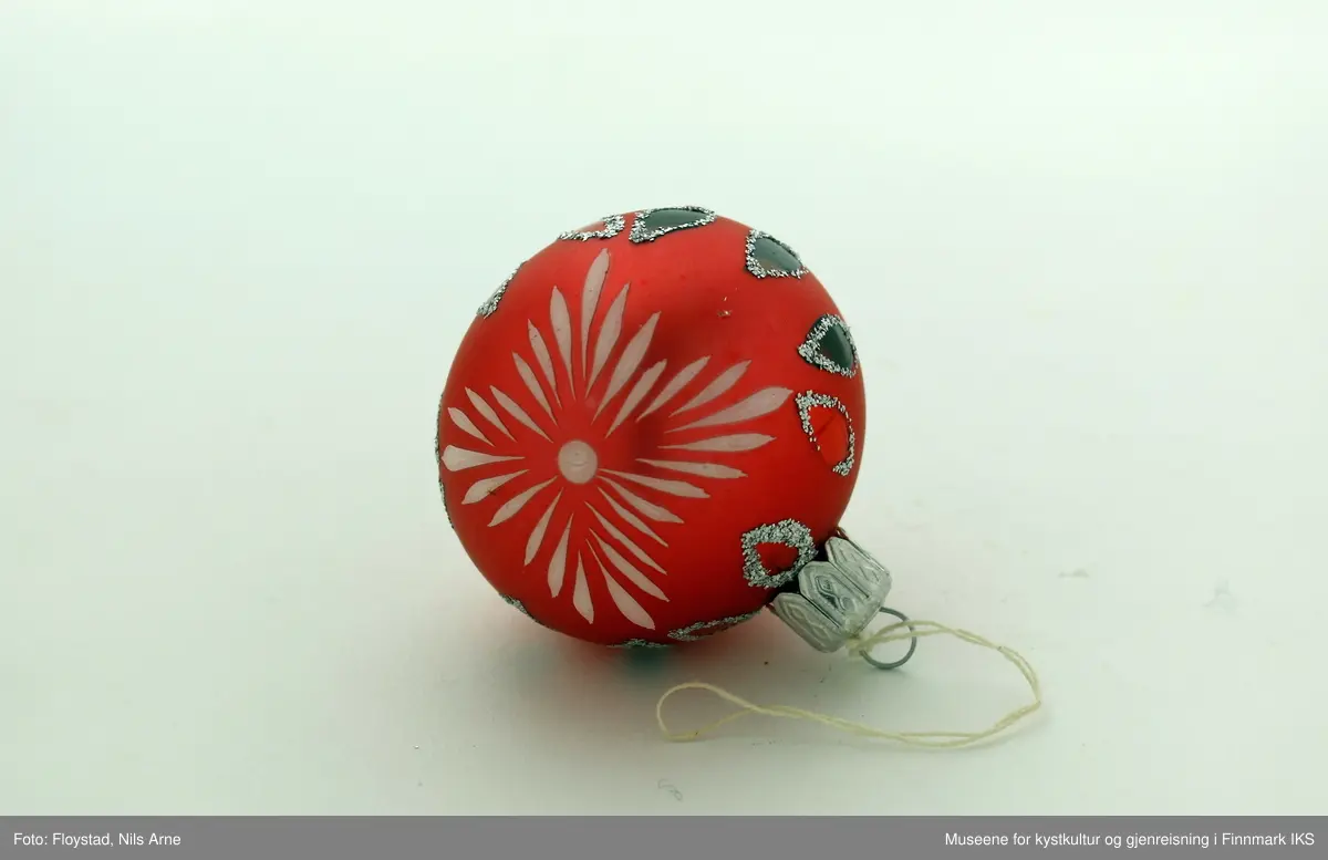 En liten julekule i hard plast med en halvmatt rød/mørkrosa farge med dekor. Dekoren består av påmalte hvite striper på begge sider av kulen og har et blomsterlignende mønster. I tillegg er det dråpeformet dekor malt rød og grønn med glitterkant som går vertikalt rundt kulen, og danner tilsammen en linje. Julekulen har et oppheng av metall med en hvit bomullstråd. Opphenget har gravert mønsterdekor.