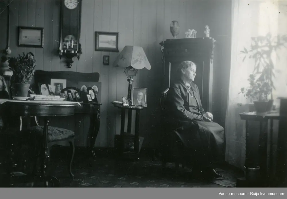 Emma Simonsen i sitt hjem på torvet i Vadsø, sommeren1937. Familieportretter står plassert i rammer på spisebordet. Emma sitter vendt mot lyset fra vinduet. På veggen henger et veggur og noen innrammede fotografier.