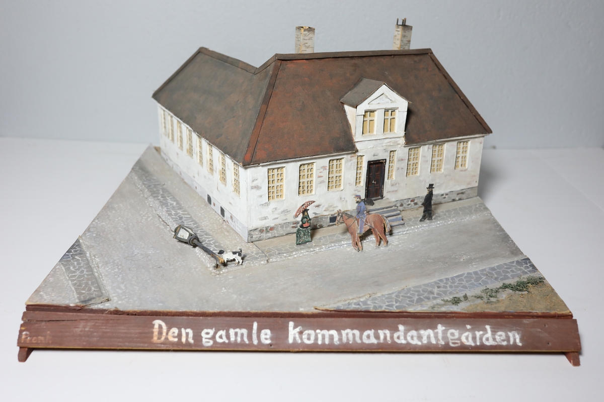 Stor rektangulær modell av den gamle kommandantgården. Modellen av bygget er satt opp på skrå slik at hjørne på huset vender mot beskueren. Baksiden av huset er umalt. Den omfatter krysset huset sto i med gatelys, hund, to personer og en mann til hest. Bak huset, i bakgården står det et tre laget av piperensere.