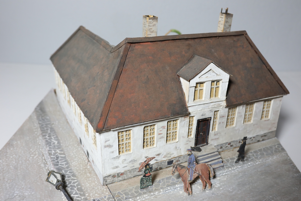 Stor rektangulær modell av den gamle kommandantgården. Modellen av bygget er satt opp på skrå slik at hjørne på huset vender mot beskueren. Baksiden av huset er umalt. Den omfatter krysset huset sto i med gatelys, hund, to personer og en mann til hest. Bak huset, i bakgården står det et tre laget av piperensere.