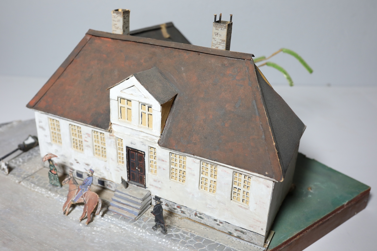 Stor rektangulær modell av den gamle kommandantgården. Modellen av bygget er satt opp på skrå slik at hjørne på huset vender mot beskueren. Baksiden av huset er umalt. Den omfatter krysset huset sto i med gatelys, hund, to personer og en mann til hest. Bak huset, i bakgården står det et tre laget av piperensere.