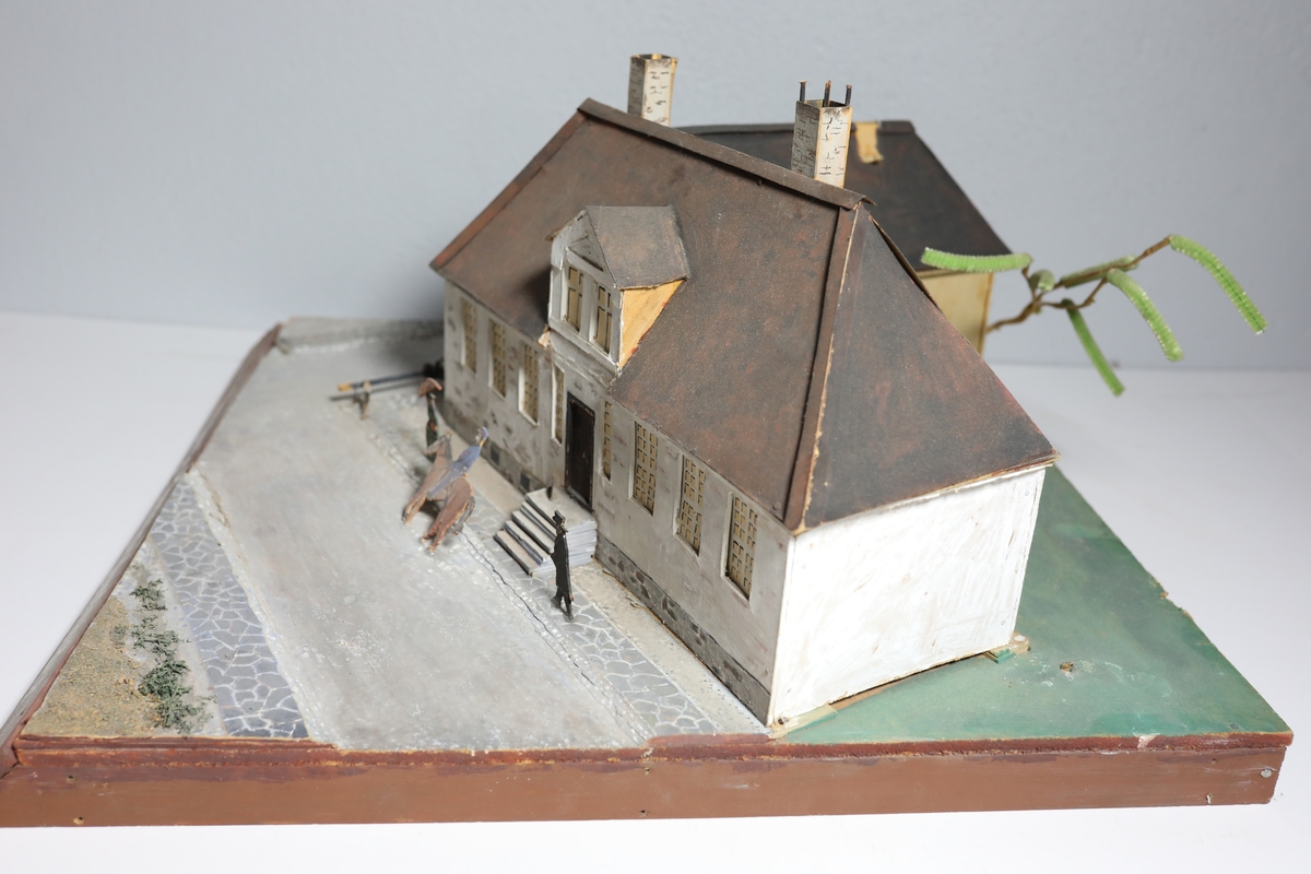 Stor rektangulær modell av den gamle kommandantgården. Modellen av bygget er satt opp på skrå slik at hjørne på huset vender mot beskueren. Baksiden av huset er umalt. Den omfatter krysset huset sto i med gatelys, hund, to personer og en mann til hest. Bak huset, i bakgården står det et tre laget av piperensere.
