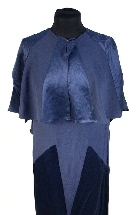 Rak aftonklänning i blått atlassiden. Ihopsydd av snedskurna kilformade våder, omväxlande på rät- och avigsida. 2 st. kilformade våder fram till midjehöjd. Pålagt stycke (med avigsidan ut) med kil 43 cm från midjan och ned. Den påsydda tygdelen är svängd och avslutad i spetsform 13 cm från halsringningen mitt fram. Bakvåden klippt i 1 stycke med kil 43 cm från midjan och ned. Rund halsringning, lite mindre bak, 0,5 cm bandkantning runt om, knäppes mitt bak med 1 tryckknapp. 20,5 cm långt sprund från halsringningen mitt bak. Ärmlös med 0,5 cm bandkantning i ärmkanten. 0,3 cm fåll nedtill på klänningen som gör kanten vågig. 
Mått i cm: längd 143, axel 1,5, midja 96.