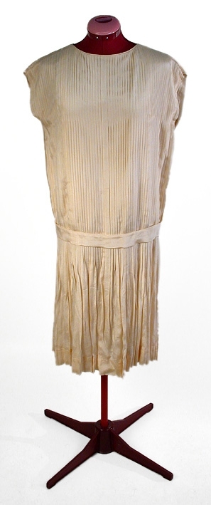 Cremefärgad, crépe de chine, helplisserad med nerhasad midja, markerad med lätt påsytt skärp (L 92 cm, B 4,5 cm). Rundad halsringning. Livet fodrat med beige siden. Ärmlös.