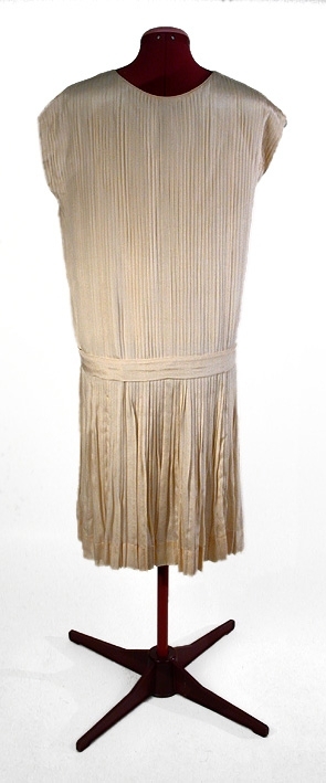 Cremefärgad, crépe de chine, helplisserad med nerhasad midja, markerad med lätt påsytt skärp (L 92 cm, B 4,5 cm). Rundad halsringning. Livet fodrat med beige siden. Ärmlös.