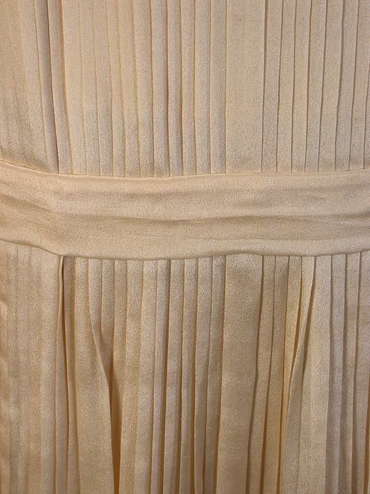 Cremefärgad, crépe de chine, helplisserad med nerhasad midja, markerad med lätt påsytt skärp (L 92 cm, B 4,5 cm). Rundad halsringning. Livet fodrat med beige siden. Ärmlös.