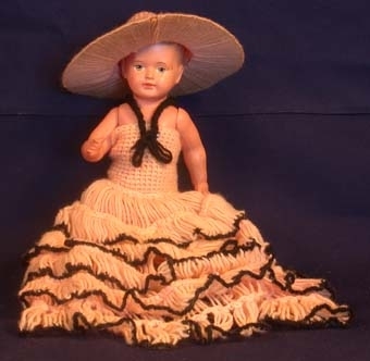 Anmärkning: Docka av beige/rosa plast med babyutseende och kort målat hår i plastrelief. Blå ögon och röda läppar med två frantänder synliga. Huvud, bröst och mage i ett stycke. Armar och ben rörliga. Dockan iklädd underbyxa (34995:2) virkad av laxrosa garn, hatt (34995:3) med brättstomme av papp runt vilken laxrosa garn virats och huvuddel av virkat laxrosa garn, samt klänning (34995:4) med volanger i kjolen av virkat laxrosa garn med svart kant runt volangerna och som axelband. Axelbandet fastsatt framtill på klänningen med knappnål. Klänningen är för stor för dockan.