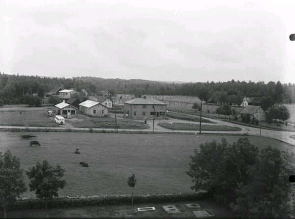 Landbygdsvy över bebyggelse kring en vägkorsning, sannolikt tagen från ett kyrktorn eftersom man ser gravar nedtill.