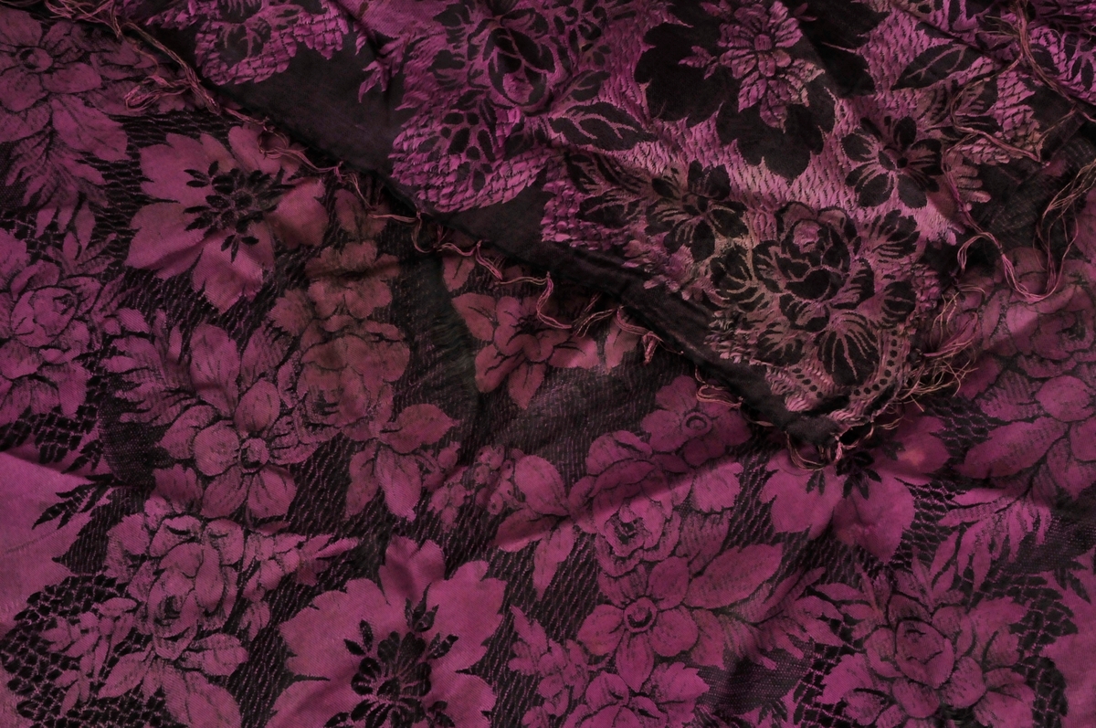 Bringeklut i mønstra silke. Tafs kring alle fire kantane. Ei mørk og ei ljosare side.