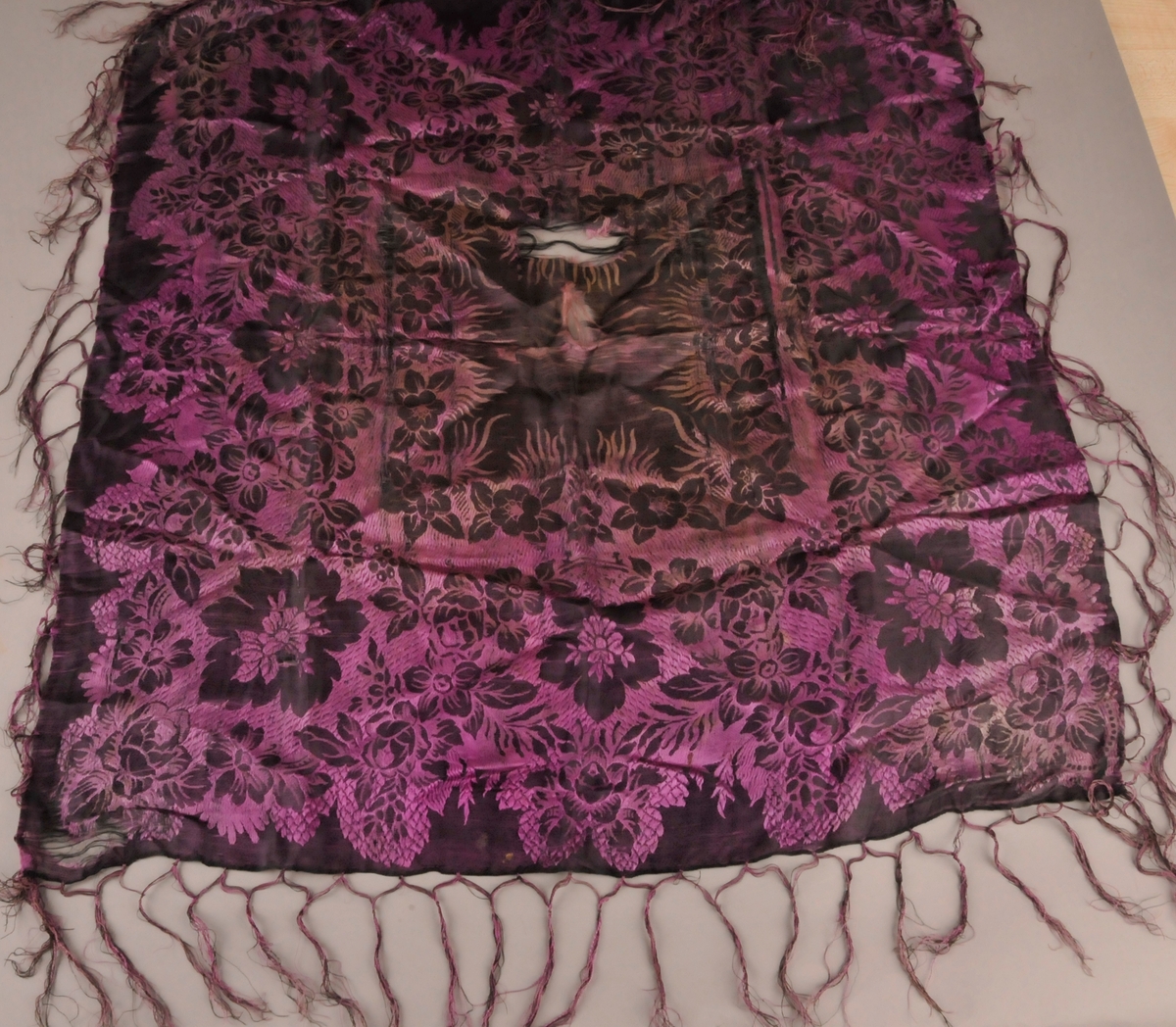 Bringeklut i mønstra silke. Tafs kring alle fire kantane. Ei mørk og ei ljosare side.