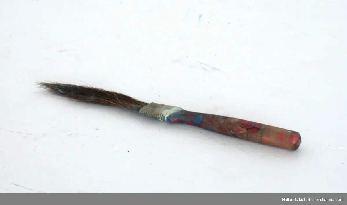Liten pensel med träskaft. Skaftet smalnar av där "penselhåren" är fastklämda med en metallbit. Blå, röd och vit målarfärg finns på skaftet.