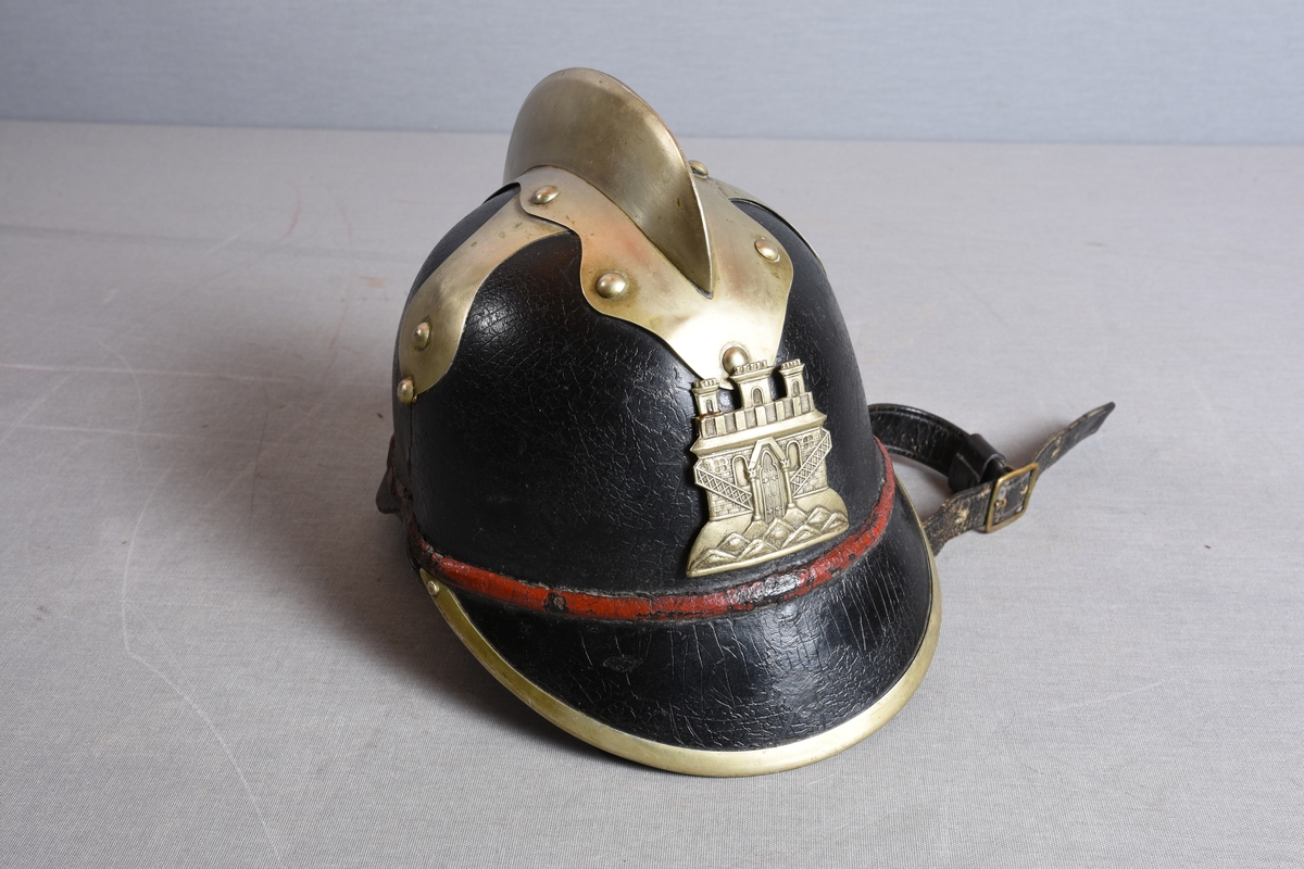 Hjelm i lær med brem foran og bak. Bremmen fremme er kantet med metall. Metallmerke i front med byvåpenmotiv. Rød stripe rundt hjelmen, like over bremmene. Hanekam på toppen festet med nagler. Fra toppen går også to metallbeslag ned på sidene. Her er noe tatt av – det er merker og hull i hjelmen som viser dette. Det kan ha vært et lignende ornament som på BBS.G00394 (lignende hjelm). Lærreim til å feste under haken. Lærforet innvendig med snorregulering.
