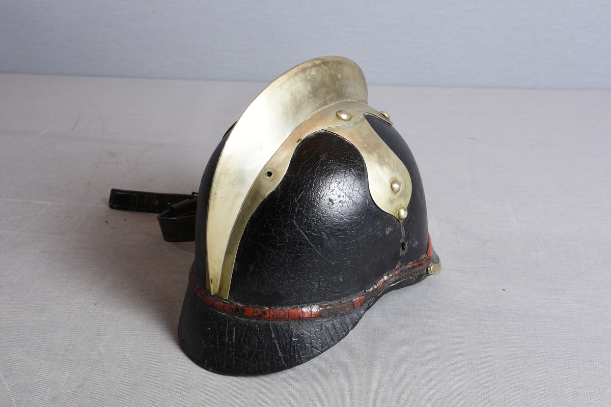 Hjelm i lær med brem foran og bak. Bremmen fremme er kantet med metall. Metallmerke i front med byvåpenmotiv. Rød stripe rundt hjelmen, like over bremmene. Hanekam på toppen festet med nagler. Fra toppen går også to metallbeslag ned på sidene. Her er noe tatt av – det er merker og hull i hjelmen som viser dette. Det kan ha vært et lignende ornament som på BBS.G00394 (lignende hjelm). Lærreim til å feste under haken. Lærforet innvendig med snorregulering.