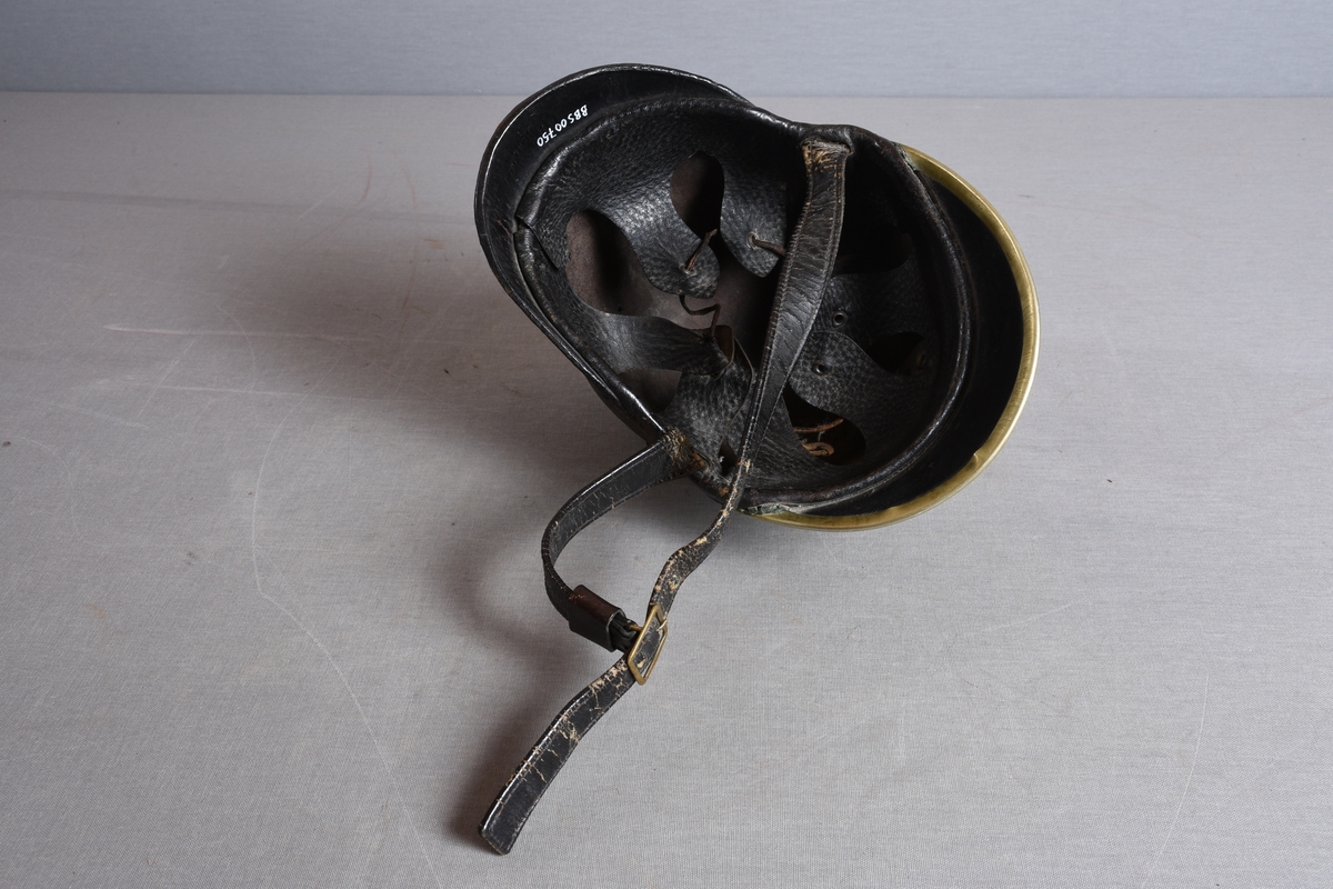 Hjelm i lær med brem foran og bak. Bremmen fremme er kantet med metall. Metallmerke i front med byvåpenmotiv. Rød stripe rundt hjelmen, like over bremmene. Hanekam på toppen festet med nagler. Fra toppen går også to metallbeslag ned på sidene. Her er noe tatt av – det er merker og hull i hjelmen som viser dette. Det kan ha vært et lignende ornament som på BBS.G00394 (lignende hjelm). Lærreim til å feste under haken. Lærforet innvendig med snorregulering.