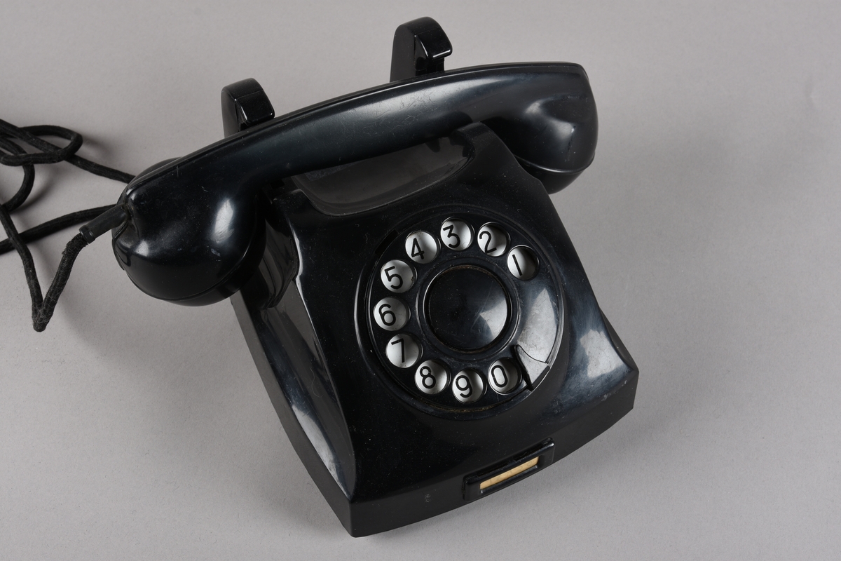 Bordtelefon med telefonrøyr og støpsel for veggkontakt. Apparatet har rektangulær grunnform med skrånande front med talskive frå 0-9. Utanpå talskiva ei skive med 10 hol der ein slår inn kvart tal i telefonnummeret ved å sette fingeren på det talet ein ønsker og dreie skiva mot høgre mot ein stoppar. På toppen av apparatet ein haldar med fjørande brytar, der kontakten vert broten når telefonrøyret ligg på haldaren. Røyret har avlang form med mikrofon i eine enden og høgtalar i andre. Leidning frå røyret til apparatet. Frå apparatet går det leidning med tre-pinna støpsel for telefonkontakt i vegg.
