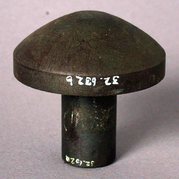 Stans och stansform (a-b). a) Stansplatta av stål. Cirkelrund med konvex profil (teckning). Längd 6,5 cm. Diameter 6,5 cm. b) Stansform av stål. Rektangulär med cylinderformad uppbyggnad i centrum. Längd 22 cm. Bredd 7,5 cm. Höjd 7 cm. 