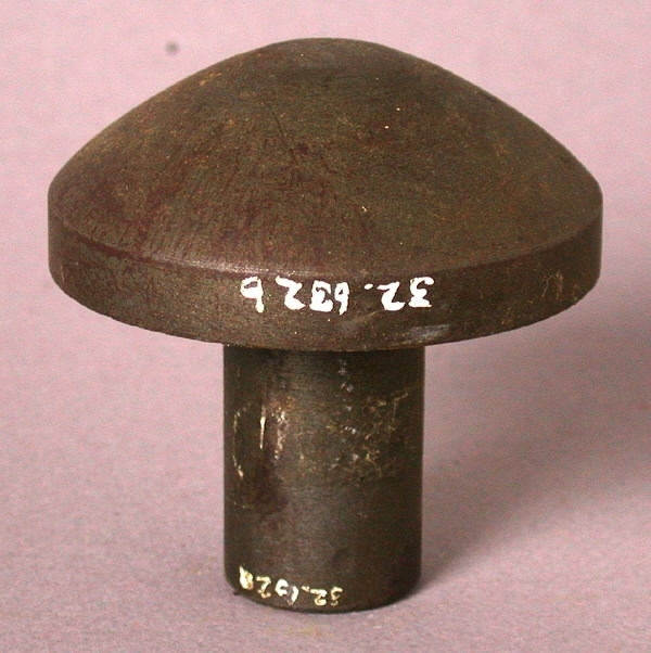 Stans och stansform (a-b). a) Stansplatta av stål. Cirkelrund med konvex profil (teckning). Längd 6,5 cm. Diameter 6,5 cm. b) Stansform av stål. Rektangulär med cylinderformad uppbyggnad i centrum. Längd 22 cm. Bredd 7,5 cm. Höjd 7 cm. 