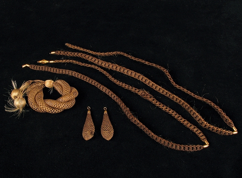 Samling bestående av fyre stycken smycken tillverkade av människohår (hårarbeten).

1: Armband av elastiskt flätat blont hår. Armbandet är försedd med en dekorativ träkula täckt av hår. I kulan har två flätade trådar fästs; trådarna har en hårkula i vardera ände. Diameter: 70 mm

2: Halsband av flätat brunt hår. Halsbandet består av två flätade delar som sammanfogats på längden med ett beslag i gulmetall. Halsbandet har ett lås av gulmetall. Längd: 580 mm

3: Halsband av flätat brunt hår. Halsbandet består av två flätade delar som sammanfogats på längden med ett beslag i gulmetall. Halsbandet har ett lås av gulmetall. Låset är skadat - en del saknas. Se bild. Längd: 580 mm

4 a-b: Örhängen av löst flätat cendrefärgat hår. Droppformade och ihåliga med beslag i gulmetall. Längd: 50mm