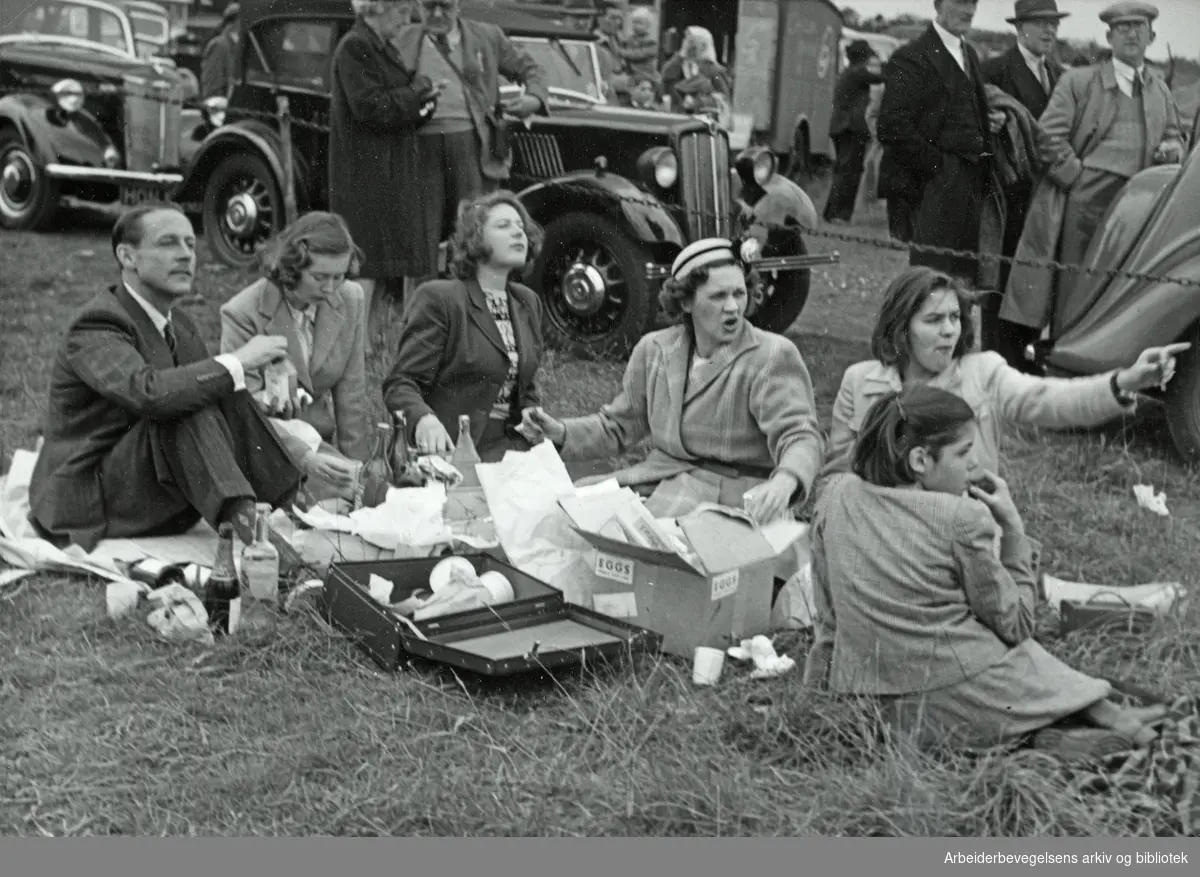 Fotoreportasje fra Derby-løpene i England. Antatt sent 1940-tall eller tidlig 1950-tall. "Denne familien nyter medbragt, men har akkurat stoppet tyggingen for å kikke på kongefamilien som nettopp gjør sin entré". Arbeidermagasinet/Magasinet for Alle