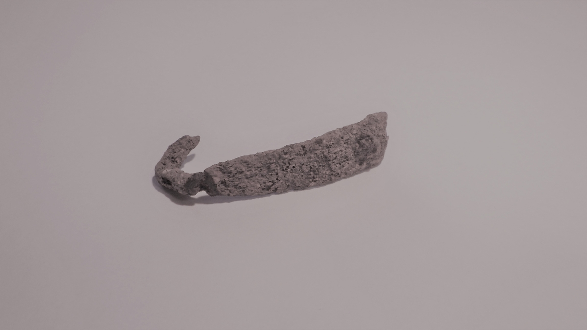 Avlang metallplate som smalner av i ene enden og som ender i en slags krok. Dette er avfall fra gjenstandsproduksjon i verkstedet.